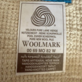 Label van een wollen handgeknoopt tapijt met het Woolmark-logo. De tekst vermeldt dat het tapijt is gemaakt van 100% zuivere scheerwol en met de hand is geknoopt.